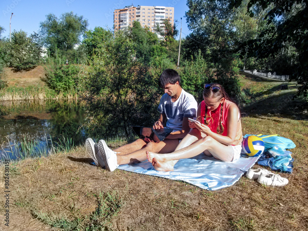 A couple of young people with gadgets are sitting on a blanket in a park near the lake. A man with a laptop and a woman with a tablet outdoors on a bright sunny summer day