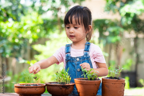 Adorable 3 years old asian little girl pointing and looking the plant  in the pots of the garden outside the house, concept of plant growing learning activity and nature education for preschool kid.