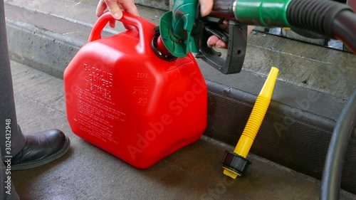 Close POV shot of a woman’s hands using a fuel pump nozzle to fill a red plastic fuel container with unleaded petrol / gas, at a gas / petrol station.