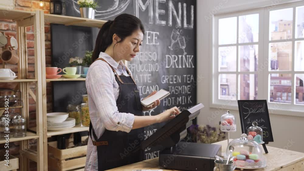 Young Asian korean girl waitress uniform making notes in notebook and typing on digital tablet on counter table. cafe female worker in apron enter customer order in point of sale terminal machine