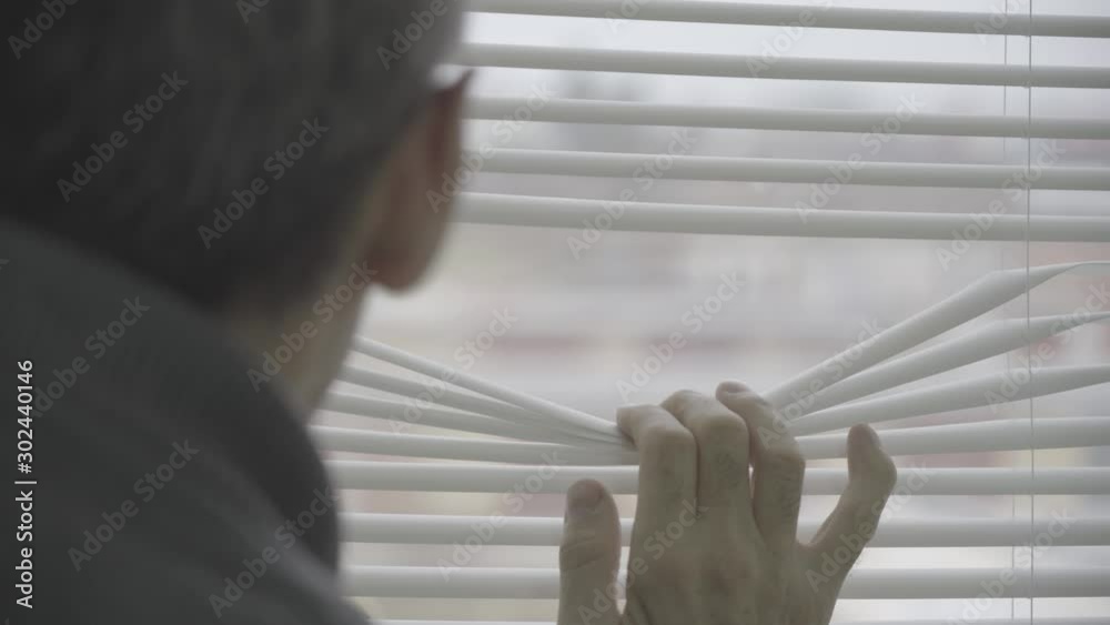The old man peers through the window. The view from the back. Selective focus on the thin fingers of a hand opening a gap in the blinds. Closeup of a mature caucasian man looking through the window bl