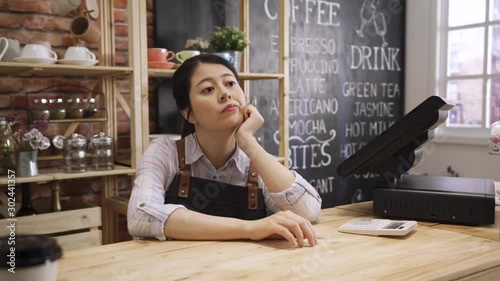Young female barman sitting leaning on wooden bar counter looking aside bored while waiting for customer in coffee shop. Beautiful asian barista in apron daydreaming with no client coming in cafe.