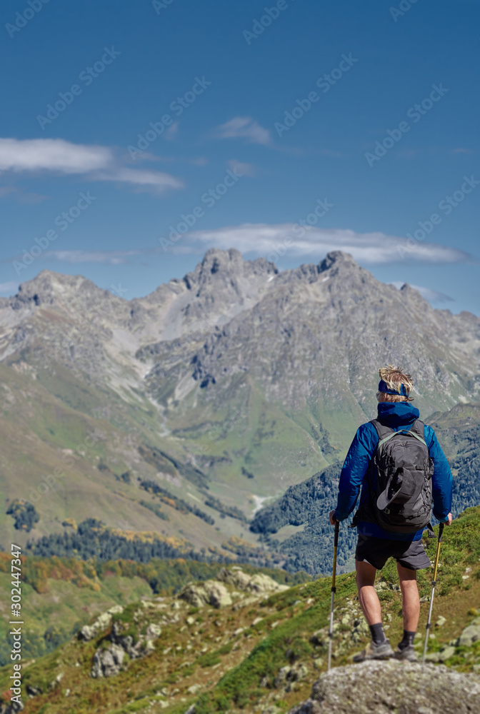 A young hiker standing on cliff edge, legs view; brave man in with climbing equipment enjoying a freedom of ascent; mountain ranges and cloudy sky on background; backpackers view from back