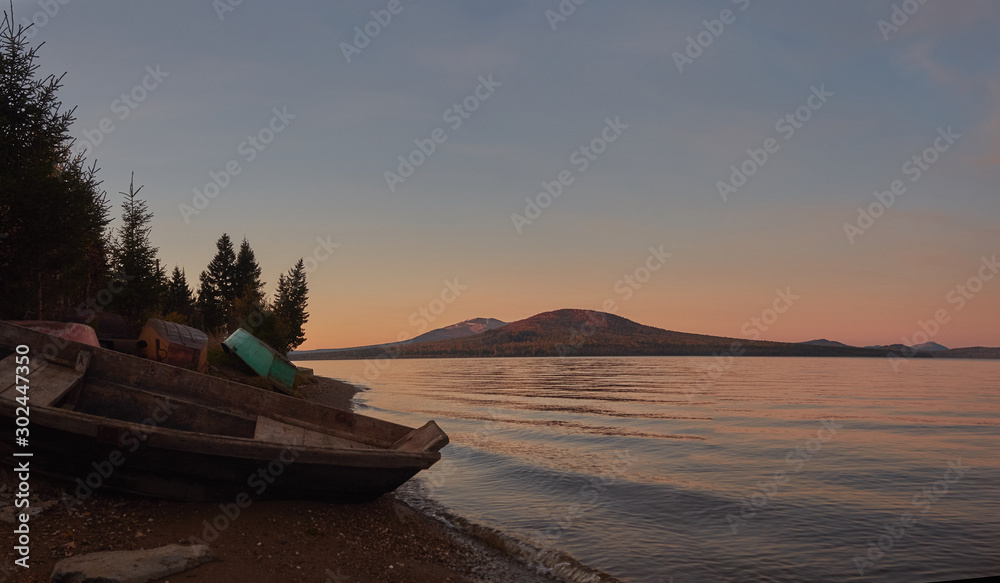 View of Zuratkul park, purple sunrise time; mountain lake coast with boats in early morning; snow tops, Ural mountain ridges on background; reflection of colorful sunrise sun on water surface View of Zuratkul park, purple sunrise time; mountain lake coast with boats in early morning; snow tops, Ural mountain ridges on background; reflection of colorful sunrise sun on water surface