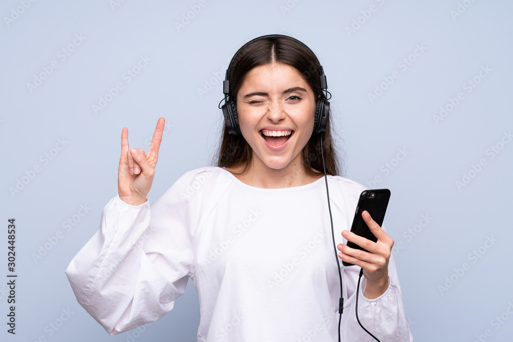 Young woman over isolated blue background using the mobile with headphones and singing Young woman over isolated blue background using the mobile with headphones and singing