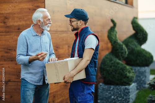 Happy delivery man talking to senior customer who is signing documents.