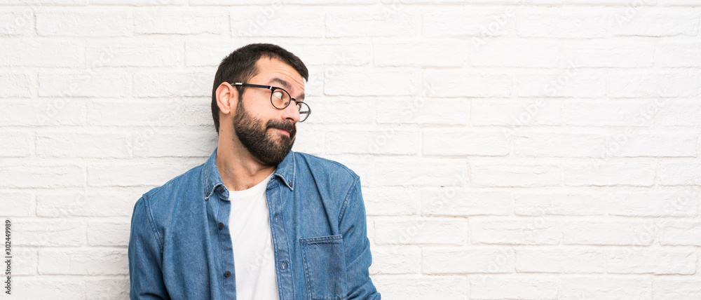Handsome man with beard over white brick wall making doubts gesture looking side Handsome man with beard over white brick wall making doubts gesture looking side