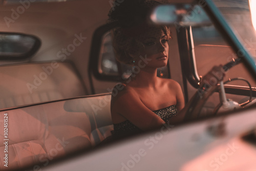 Portrait of beautiful sexy fashion blond girl. Model with bright makeup and curly hairstyle in retro style hat sitting in old car. Attractive woman  rides around the night city