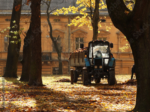 Tractor among the trees in the autumn park of the city of Lviv. The municipal service of the city removes fallen leaves. Change of seasons. Wilting nature. An abundance of warm colors. Fiery colors