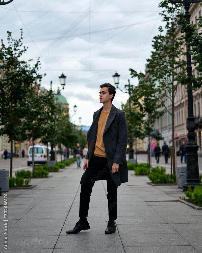 Handsome stylish fashionable man, brunette in elegant gray coat, stands on street in historical center of St. Petersburg. Young man with dark hair, thick eyebrows. Handsome stylish fashionable man, brunette in elegant gray coat, stands on street in historical center of St. Petersburg. Young man with dark hair, thick eyebrows.