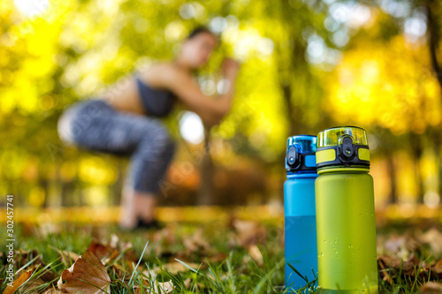 two bottles of water on green grass. sporty woman doing outdoor workout in park on background. focus on bottle