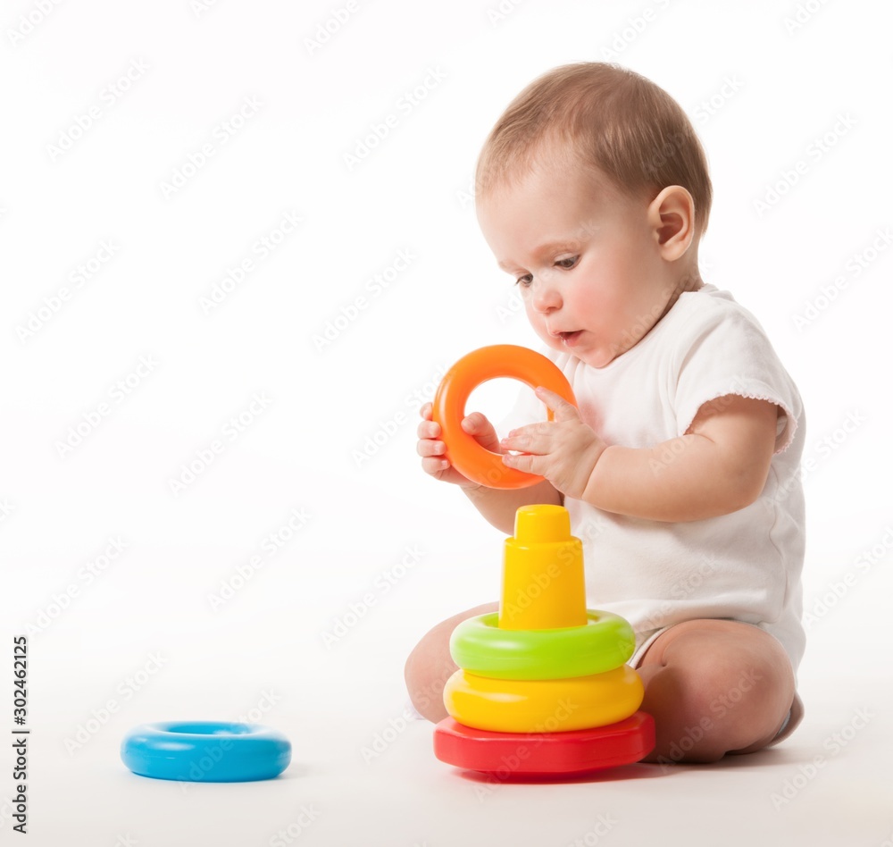 Charming cute baby in white overalls holds rings of a colored plastic pyramid sitting on a white background. Concept of smart and healthy little children concept of child care. Advertising space Charming cute baby in white overalls holds rings of a colored plastic pyramid sitting on a white background. Concept of smart and healthy little children concept of child care. Advertising space