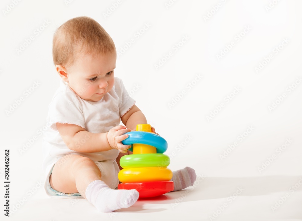 Charming cute baby in white overalls holds rings of a colored plastic pyramid sitting on a white background. Concept of smart and healthy little children concept of child care. Advertising space Charming cute baby in white overalls holds rings of a colored plastic pyramid sitting on a white background. Concept of smart and healthy little children concept of child care. Advertising space