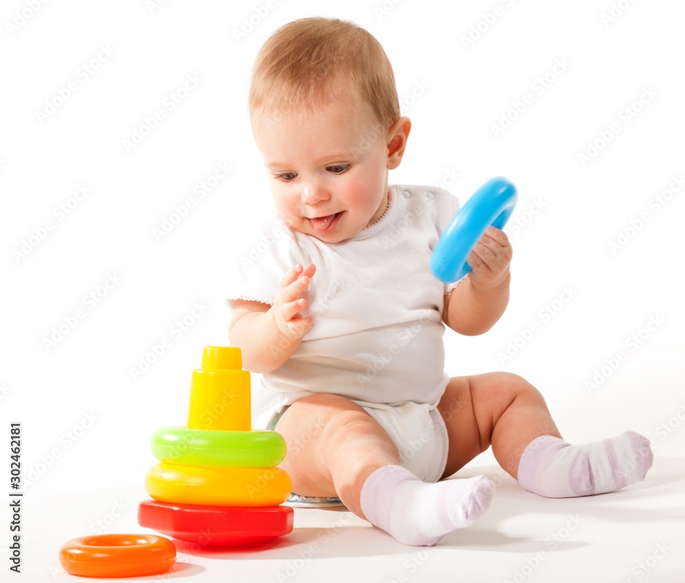 Charming cute baby in white overalls holds rings of a colored plastic pyramid sitting on a white background. Concept of smart and healthy little children concept of child care. Advertising space Charming cute baby in white overalls holds rings of a colored plastic pyramid sitting on a white background. Concept of smart and healthy little children concept of child care. Advertising space