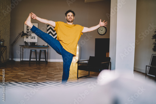 Experienced young man standing on one leg and stretching another during yoga to develop flexibility and achieve harmony of body.Hipster guy engaged in meditation and practicing healthy lifestyle