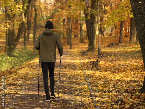 A young guy is engaged in Nordic walking in an autumn park among fallen yellow foliage. The physical activity of a healthy lifestyle. Change of seasons. Wilted nature. An abundance of warm colors