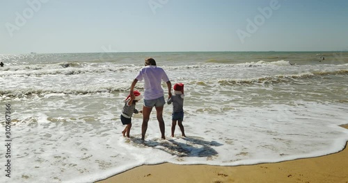 young mother holds pretty small daughters wearing red hats and runs across sand beach to sea water edge against warm waves