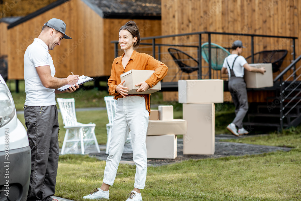 Couriers delivering goods to a young woman home by cargo van vehicle, client signing documents, mover with cardboard parcels on the background