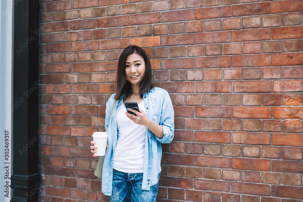 Cheerful Asian lady next to wall messaging on smartphone Cheerful Asian lady next to wall messaging on smartphone