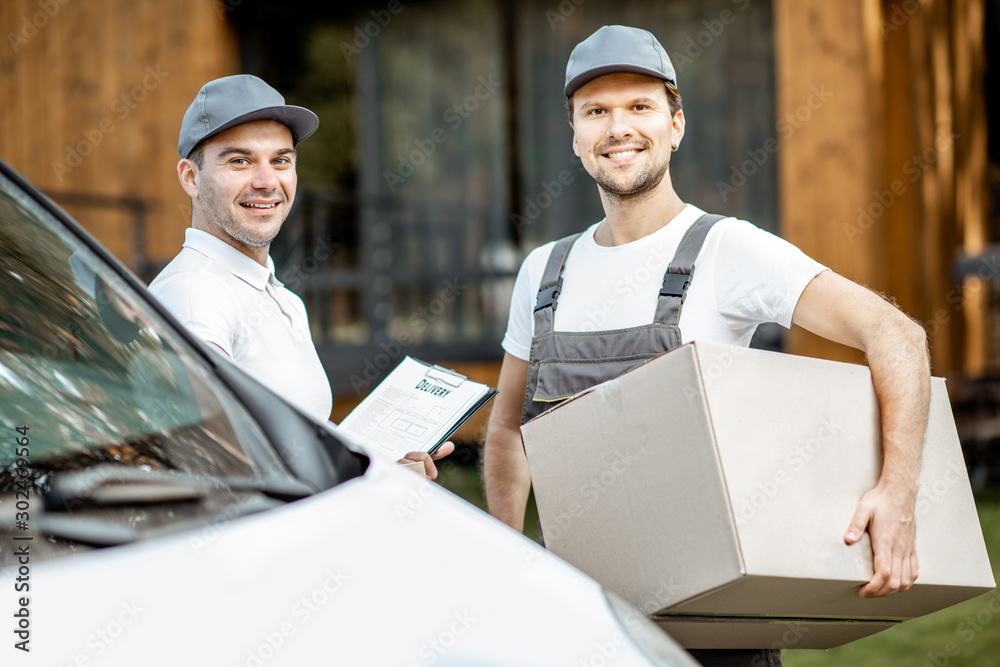 Portrait of a two cheerful delivery men in uniform standing together with check list and cardboard boxes near the cargo van vehicle outdoors Portrait of a two cheerful delivery men in uniform standing together with check list and cardboard boxes near the cargo van vehicle outdoors