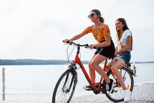 Two female friends on the bike have fun at beach near the lake
