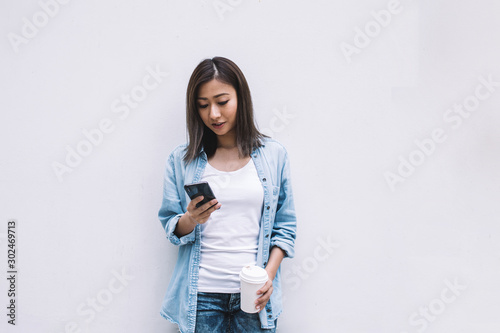 Young beautiful Asian woman leaning on wall checking smartphone with takeaway coffee photo