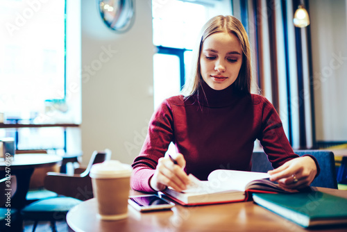 Young attractive woman blonde dressed in casual clothes planning working schedule while sitting in cozy coffee shop.Charming skilled female student preparing for examination using textbook indoors