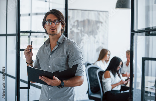 Thoughtful look. Man in glasses with notepad and pen in hands stands in the office opposite employees