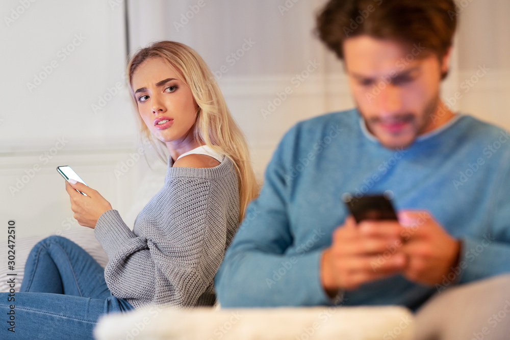 Wife Watching Husband Chatting On Cellphone Sitting On Couch Indoor Wife Watching Husband Chatting On Cellphone Sitting On Couch Indoor