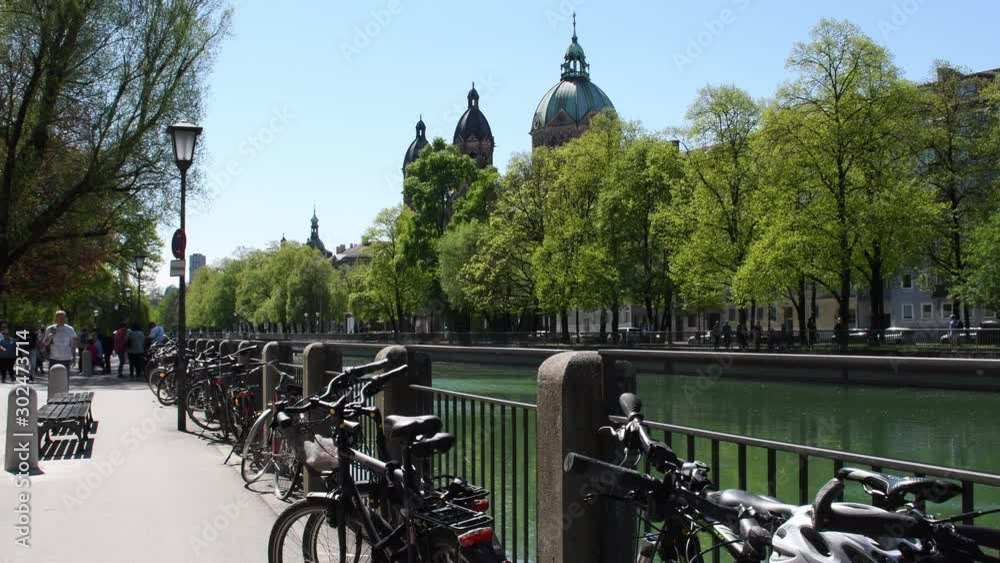 Many bicycles stand on the embankment of the Isar River in Munich. People are walking along the promenade on a warm Easter day, green trees and a St. Lukas Church are in the background.