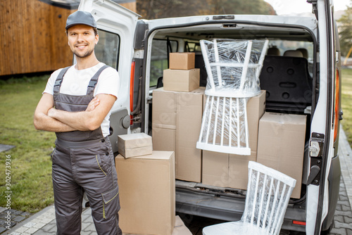 Portrait of a handsome delivery man in uniform standing near a cargo van vehicle trunk full of boxes and furniture during a relocation