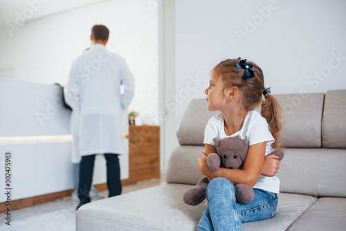 Doctors behind. Cute little girl with teddy bear in hands sits in waiting room of hospital