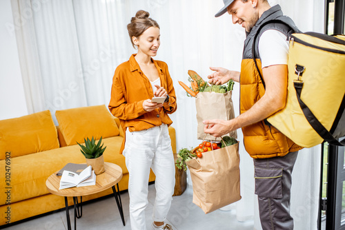 Courier in uniform with thermal bag delivering fresh groceries in paper bags to a client home. Woman with smartphone checking her order photo