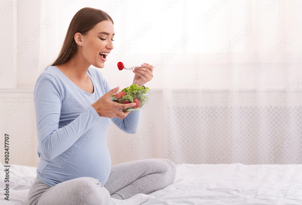 Pregnant Girl Eating Vegetable Salad Sitting On Bed In Bedroom