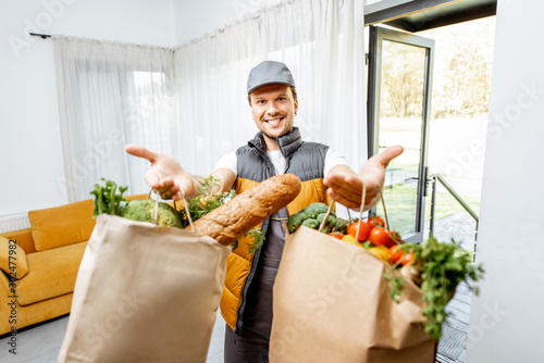 Portrait of a cheerful courier in uniform delivering fresh groceries home, holding paper bags full of food in front of the camera indoors photo