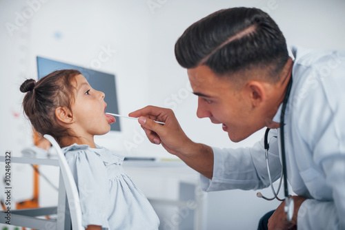 Uses tonsil to check throat. Young pediatrician works with little female visitor in the clinic photo