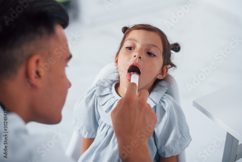 Uses tonsil to check throat. Young pediatrician works with little female visitor in the clinic
