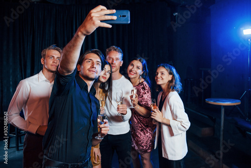 Takes selfie. Group of cheerful friends celebrating new year indoors with drinks in hands