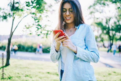 Cheerful hipster girl in eyewear downloading song to smartphone playlist while spending spring day in park, smiling young woman connecting to radio broadcast using mobile phone and earphones on leisur photo