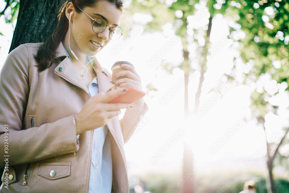 Attractive hipster girl in eyewear checking notification on smartphone download new song to playlist, female meloman listening to music in earphones via app while having coffee break outdoors