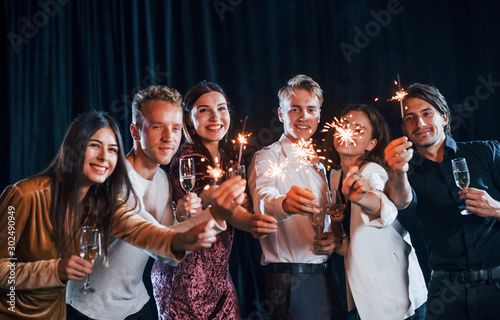 Having fun with sparklers. Group of cheerful friends celebrating new year indoors with drinks in hands