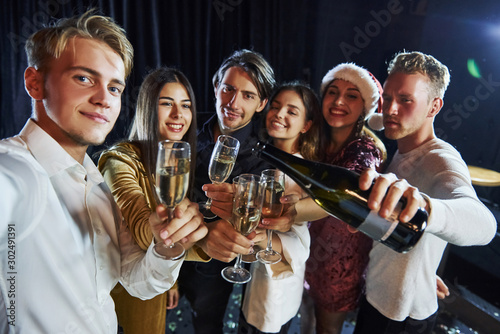 Takes selfie. Group of cheerful friends celebrating new year indoors with drinks in hands