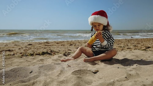 funny girl celebrates Christmas sitting on tropical sand beach eating yellow corn and sneezing at calm ocean slow motion photo