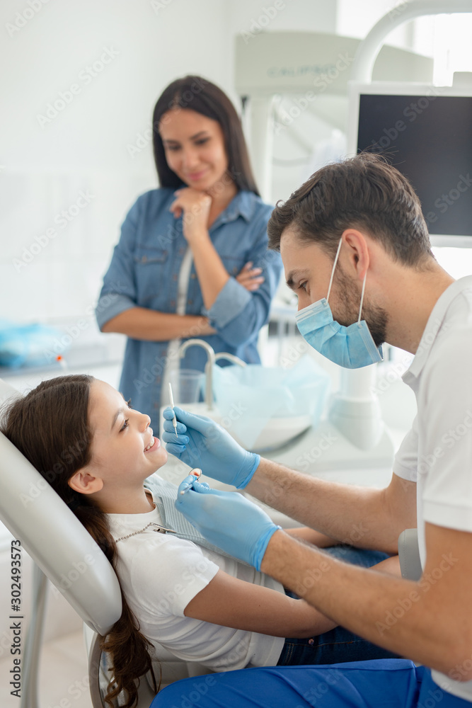 male doctor with instruments in the hands looking girls teeth sitting in the dental chair while mother standing near her for support male doctor with instruments in the hands looking girls teeth sitting in the dental chair while mother standing near her for support