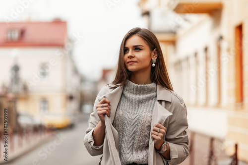 Pretty cute young woman with chic brown long hair with natural make-up in fashionable trench coat in knitted sweater posing in a spring day outdoors. Stylish attractive girl model resting in the city.