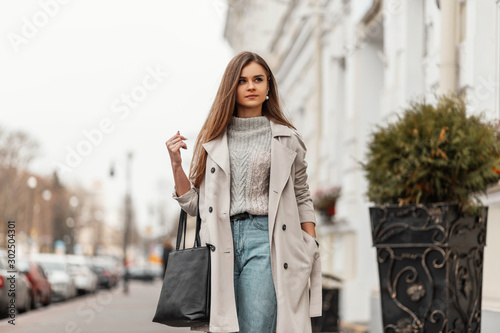 Photos Pretty trendy young woman in a stylish trench coat in a vintage knitted sweater in blue jeans with a leather bag enjoys a walk along the street
