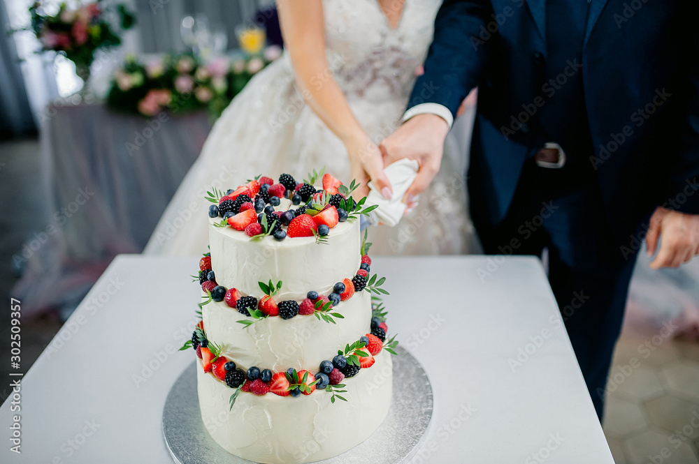 wedding ceremony. Hands of newlyweds cut a white three-tiered cake with strawberries and blackberries wedding ceremony. Hands of newlyweds cut a white three-tiered cake with strawberries and blackberries