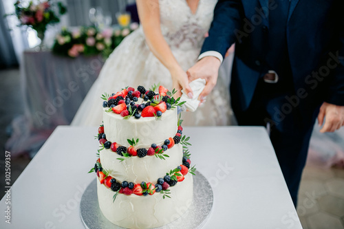 wedding ceremony. Hands of newlyweds cut a white three-tiered cake with strawberries and blackberries