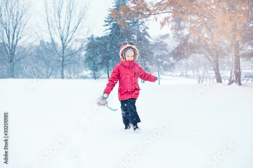 Cute adorable funny Caucasian smiling laughing girl child in warm clothes red pink jacket running in snow and having fun during cold winter snowy day. Kids outdoor seasonal activity.