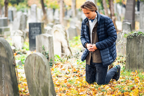 Mourning young man kneeling in front of a grave on a cemetery